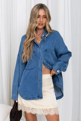 A woman wearing an oversized blue denim button-up shirt over a cream lace-trim mini skirt, standing against a white curtain background.