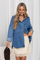 A woman wearing an oversized blue denim button-up shirt over a cream-colored skirt, holding a dark brown textured handbag against a white curtain background.