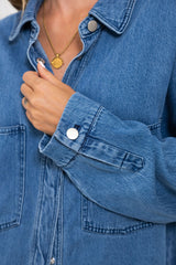 Close-up of a person wearing a medium-wash blue denim button-up shirt jacket, showing the chest area, collar, front pockets, and a hand adjusting the collar.