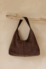 A dark brown suede hobo shoulder bag hanging from a natural wood branch against a textured beige plaster wall.