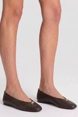A close-up of a model's feet wearing dark brown leather ballet flats with a small gold-tone metal accent on the toe.