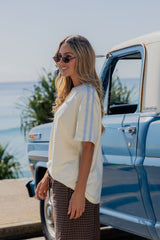 A woman wearing a cream short-sleeve t-shirt with light blue stripes on the sleeves and brown plaid pants, standing next to a vintage blue truck with the ocean in the background.