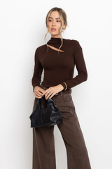 Woman wearing a brown long-sleeve top and pants holding a black handbag against a white background