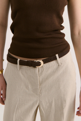 A close-up of a person wearing a dark brown ribbed knit top, a dark brown belt with a gold-tone oval buckle, and light beige trousers against a plain light background.