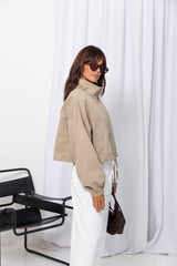 Woman wearing a stone funnel neck bomber jacket over white pants, styled with a brown suede bag, on a white background.