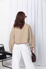 Woman wearing a stone funnel neck bomber jacket over white pants, styled with a brown suede bag, on a white background.