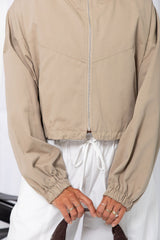 Woman wearing a stone funnel neck bomber jacket over white pants, styled with a brown suede bag, on a white background.