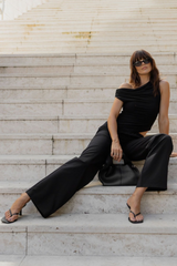 A woman wearing a black off-the-shoulder top and matching wide-leg pants sits on light stone steps, with a black handbag beside her.