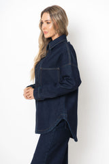 A woman wearing an oversized dark indigo denim shirt jacket with a pointed collar and front chest pocket, shown in profile against a plain white studio background.