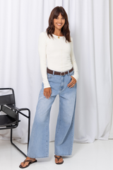 Woman wearing a white long-sleeve top with light-wash wide-leg jeans and a brown belt, carrying a brown handbag, on a white background.