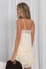 A woman shown from the back wearing a cream satin slip dress with lace trim, standing against a white curtain background and holding a dark brown knit bag.