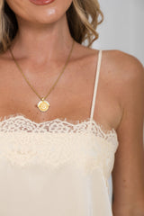 A close-up of a model wearing a delicate gold chain necklace with a round, textured gold pendant, styled with an ivory lace-trimmed camisole top.