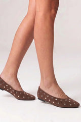 A close-up of a person's legs wearing brown pointed-toe flats decorated with small silver studs, set against a plain light gray background.
