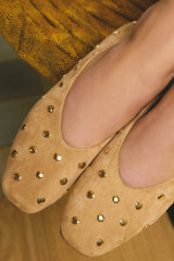 Close-up of a person wearing tan suede ballet flats decorated with small gold-tone studs, with a mustard yellow textured garment visible in the background.