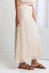A model wearing a midi-length beige crochet skirt with a ribbed elastic waistband, in a fitted silhouette, paired with a crochet top and brown sandals.