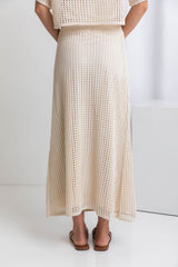 A model wearing a midi-length beige crochet skirt with a ribbed elastic waistband, in a fitted silhouette, paired with a crochet top and brown sandals.