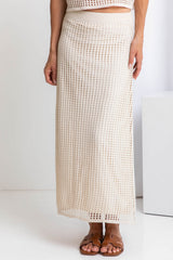 A model wearing a midi-length beige crochet skirt with a ribbed elastic waistband, in a fitted silhouette, paired with a crochet top and brown sandals.