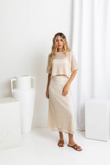 A model wearing a crochet top and midi-length beige crochet skirt with a ribbed elastic waistband, in a fitted silhouette, paired with a crochet top and brown sandals.