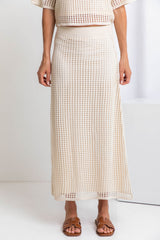 A model wearing a midi-length beige crochet skirt with a ribbed elastic waistband, in a fitted silhouette, paired with a crochet top and brown sandals.