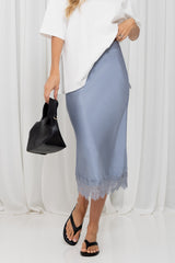 A model wearing a light blue satin midi skirt with a lace hem, styled with a white top, black sandals, and holding a black handbag against a white curtain backdrop.