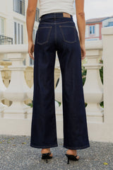 A person shown from the back wearing dark indigo wide-leg jeans, a white ribbed top, and black high-heeled sandals, standing outdoors in front of a white balustrade railing.