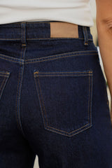 Close-up of the back waistband and back pocket of dark indigo denim jeans, worn with a white ribbed top. A tan leather brand patch is visible at the back waistband.