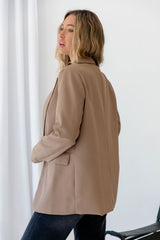 A woman wearing an oversized taupe blazer and dark jeans, photographed from the back and side against a bright white indoor background with a curtain.