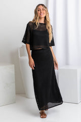 A woman wearing a black crochet knit crop top with a scoop neckline and 1/4 sleeve, paired with a black skirt.