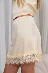 A close-up of a model wearing a cream satin mini skirt with a scalloped lace hem, paired with a matching cream satin top.