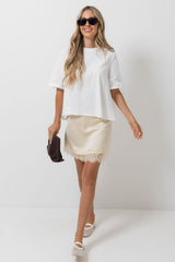 A woman modeling a white short-sleeve crop top and a cream satin mini skirt with lace trim, styled with black sunglasses, a dark brown clutch, and white platform sandals against a plain white studio background.