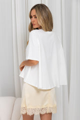 A woman wearing a white short-sleeve cape-style top and a cream satin skirt with a lace hem, photographed indoors against white curtains.