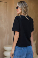 A woman with blonde hair and sunglasses, seen from behind, wearing a black short-sleeve t-shirt and blue jeans in a warm-toned indoor setting.