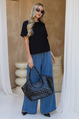 A woman wearing a black t-shirt, wide-leg blue jeans, and black sunglasses holds a black patterned tote bag in a modern indoor setting with beige walls and white drapes.