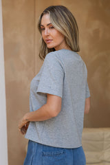 A woman with blonde-highlighted shoulder-length hair is shown from the back wearing a light heather gray short-sleeve crop top and blue jeans, standing against a warm beige textured wall.