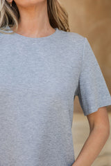 A close-up of a woman wearing a light heather gray short-sleeve crewneck t-shirt, cropped from the chin down against a warm beige background.