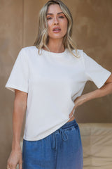A woman wearing a white short-sleeve crewneck t-shirt and blue denim drawstring pants, standing against a textured beige wall.