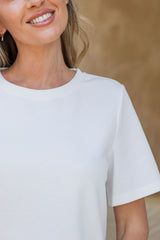 A close-up of a woman wearing a plain white short-sleeve crewneck t-shirt, cropped from the mouth down against a warm beige textured background.