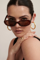 A close-up of a model wearing dark brown cat-eye sunglasses with gold hoop earrings and a gold ring, set against a plain light beige background.