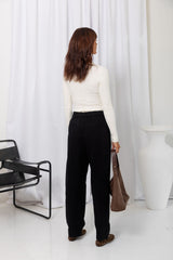 Woman wearing a white long-sleeve top with black tailored trousers, carrying a brown suede bag, on a white background.