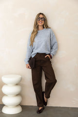 Woman wearing a gray sweater and brown pants standing against a beige wall.