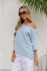 A woman wearing a light blue off-the-shoulder knit top and white pants, standing against a white wall with green palm leaves in the background.