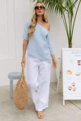 A woman wearing a light blue off-the-shoulder knit top and white wide-leg pants, carrying a woven straw tote bag. She is standing in a bright, modern setting with a white wall, a potted palm, and a small gray stool.