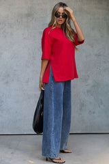 A woman modeling a bright red short-sleeve top with side slits, paired with wide-leg blue jeans, black sunglasses, and carrying a black tote bag against a textured gray concrete wall.