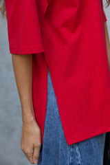 A close-up of a person wearing a bright red oversized short-sleeve top with a side slit, paired with blue jeans, against a gray concrete wall.