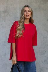 A woman with blonde wavy hair wearing a bright red oversized short-sleeve top and blue jeans, standing against a textured gray concrete wall.