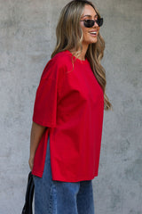 A woman wearing an oversized bright red short-sleeve top and blue jeans, standing against a textured gray concrete wall.