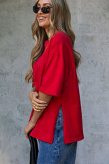 A woman wearing an oversized bright red short-sleeve top and blue jeans, posing against a textured gray concrete wall.