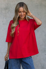 A woman wearing a bright red oversized short-sleeve t-shirt and blue jeans, standing against a textured gray concrete wall.