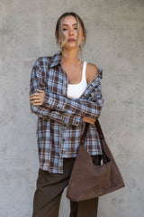 Woman wearing a plaid shirt over a white top, holding a brown leather bag against a concrete wall.