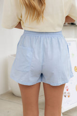 A person shown from the back wearing light blue shorts with an elastic waistband and side pockets, paired with a cream top in a bright indoor setting.